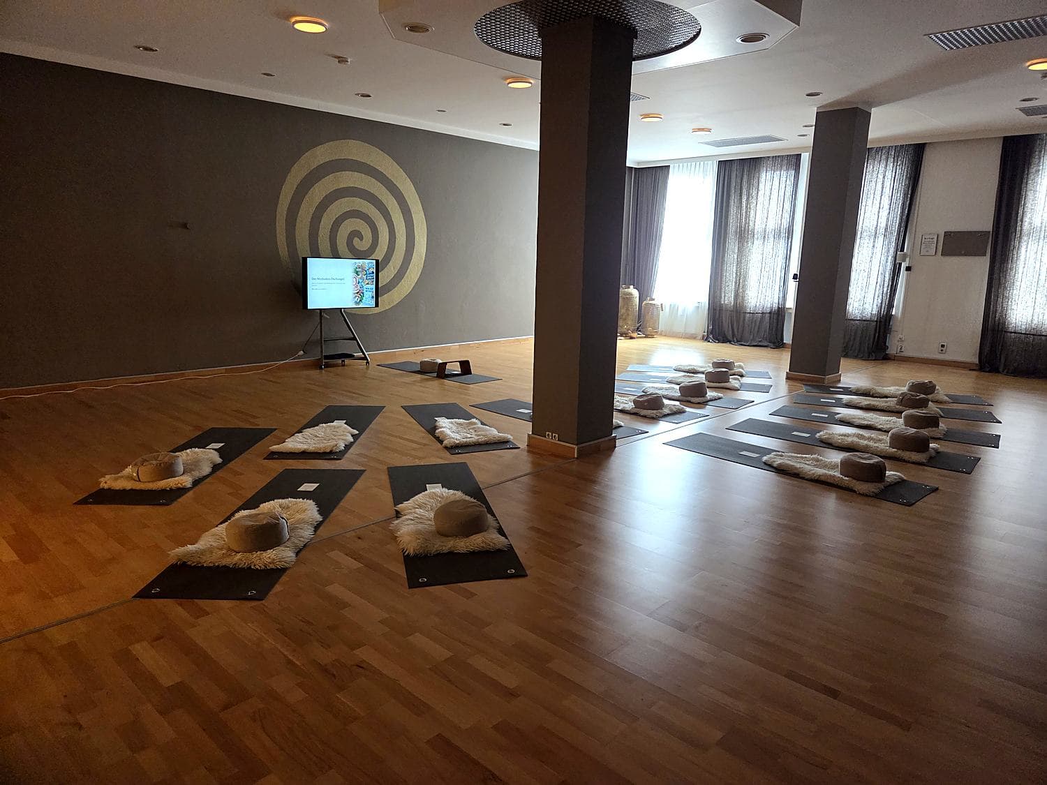 Ein Seminarraum mit ausgelegten Matten, Schaffellen und Sitzkissen, vorne ein Bildschirm vor einer Wand mit goldener Spirale