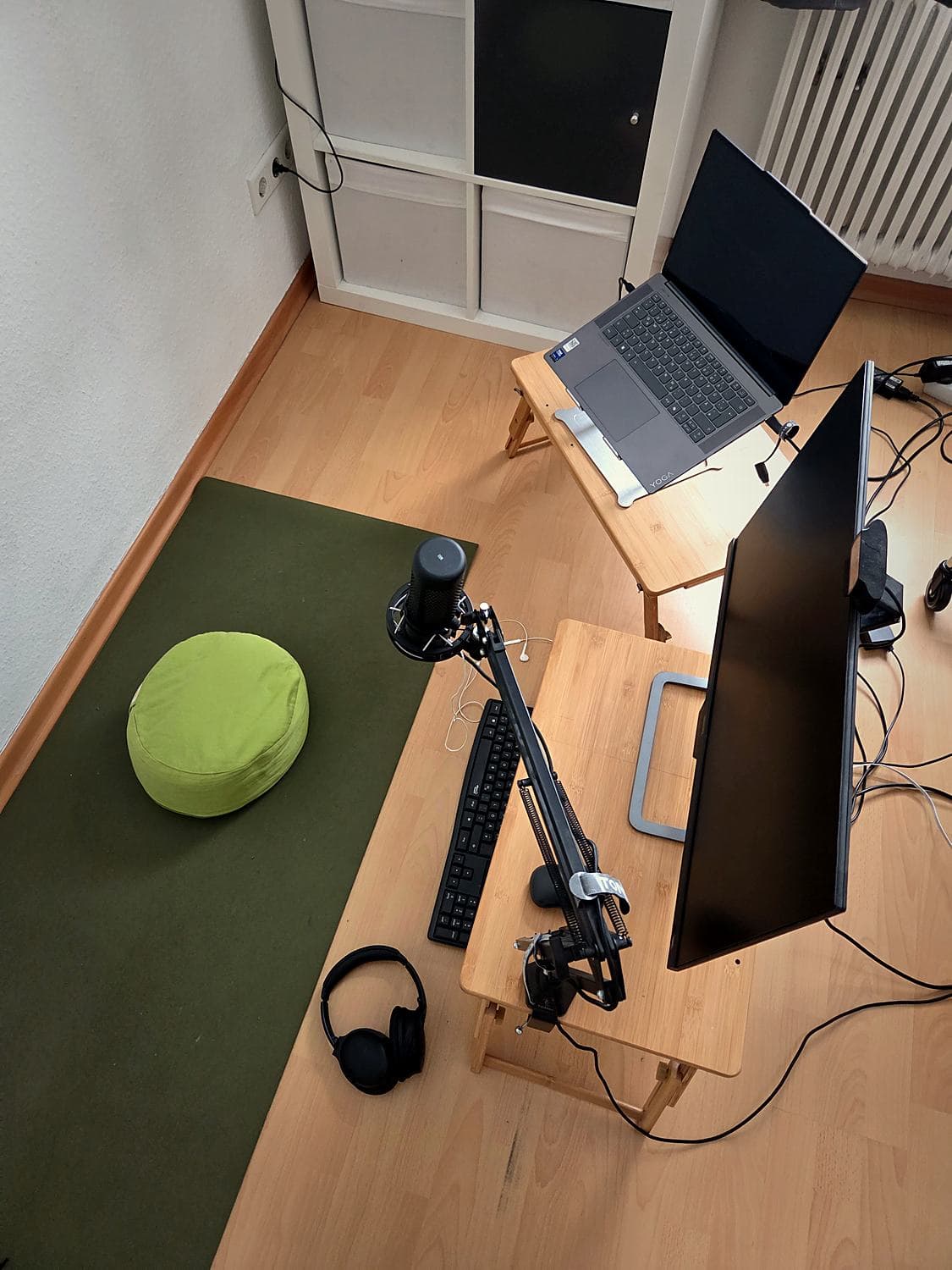 Blick von oben auf einen Schreibtisch mit Mikrofon am Schwenkarm, Tastatur, Kopfhörern und Monitor, daneben eine Matte mit grünem Sitzkissen
