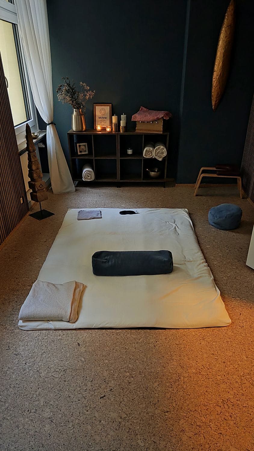 Eine bodentiefe Liegematte mit hellem Laken, dunklem Bolster, Decke und Augenkissen, dahinter ein Regal mit gerollten Handtüchern, Klangschale und Kerzen