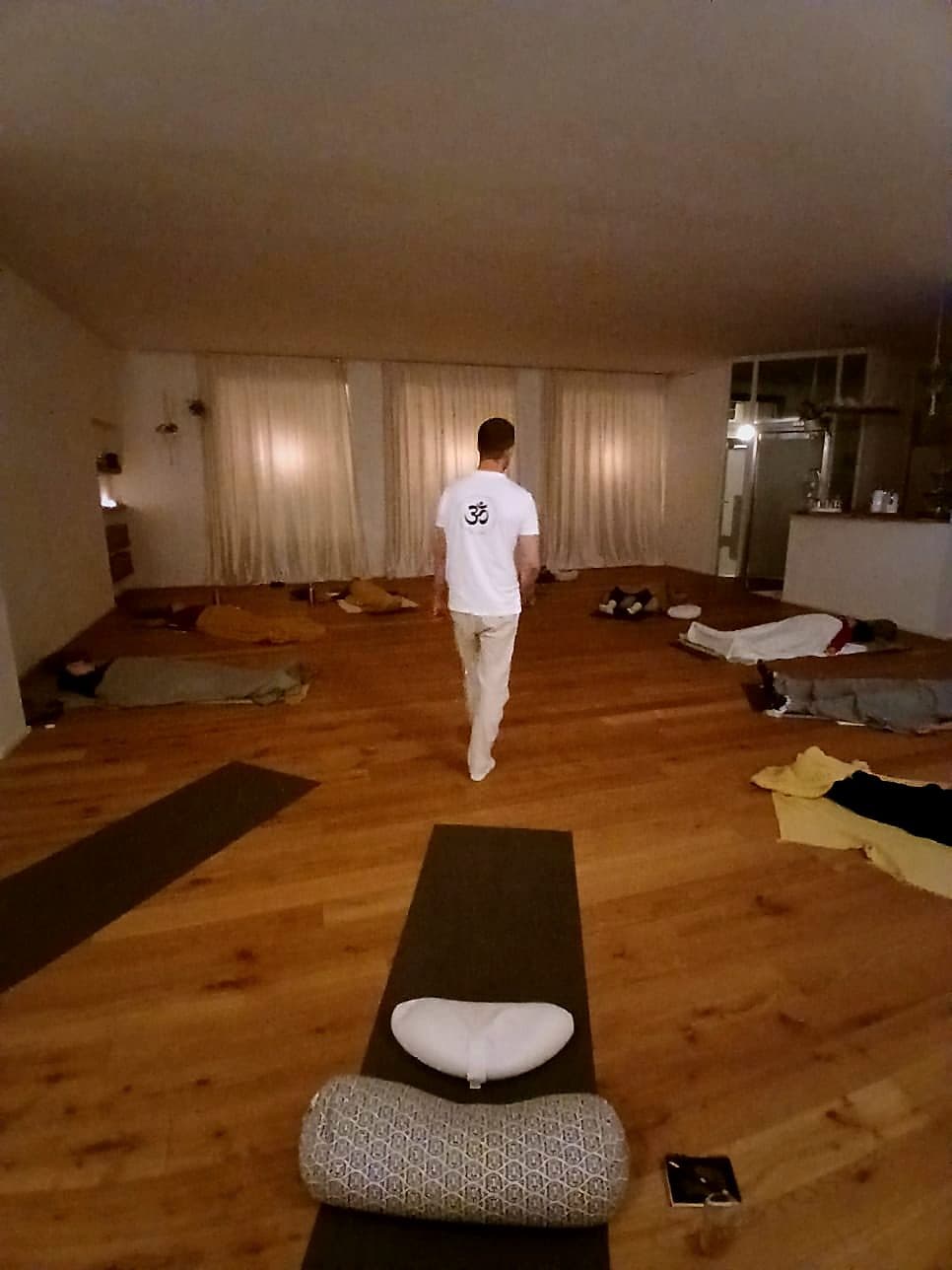 Nils Hammann geht in weißem Shirt durch einen Raum, in dem ringsum Menschen unter Decken auf Matten liegen