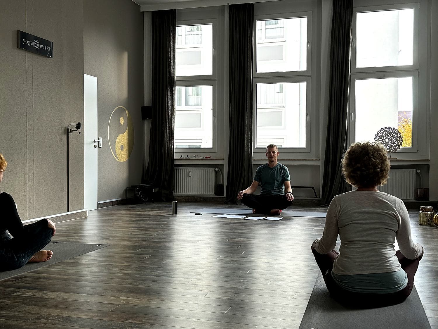 Nils Hammann sitzt im Schneidersitz vor zwei Teilnehmerinnen in einem hellen Studio mit hohen Fenstern und goldenem Yin-Yang-Zeichen an der Wand